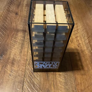 Peut inclure: Une tour de jeu en plastique transparent avec des blocs de bois. Le jeu s'appelle STAR ATTACK, avec le logo visible &agrave; l'avant. Les blocs sont de couleur marron clair et empil&eacute;s &agrave; l'int&eacute;rieur de la tour. La tour est pos&eacute;e sur une surface en bois.