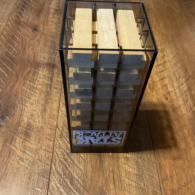 Peut inclure: Une tour de jeu en plastique transparent avec des blocs de bois. Le jeu s'appelle STAR ATTACK, avec le logo visible &agrave; l'avant. Les blocs sont de couleur marron clair et empil&eacute;s &agrave; l'int&eacute;rieur de la tour. La tour est pos&eacute;e sur une surface en bois.