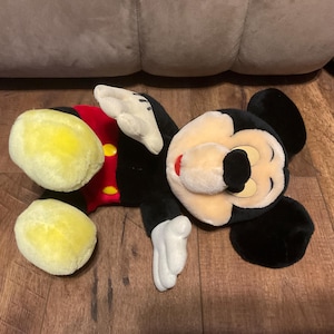 Pu&ograve; includere: Un peluche di Topolino, sdraiato su una superficie di legno. Il giocattolo presenta un corpo nero, grandi piedi gialli, guanti bianchi e una camicia rossa con bottoni gialli. Il viso &egrave; color crema con un naso nero e un'espressione sorridente.