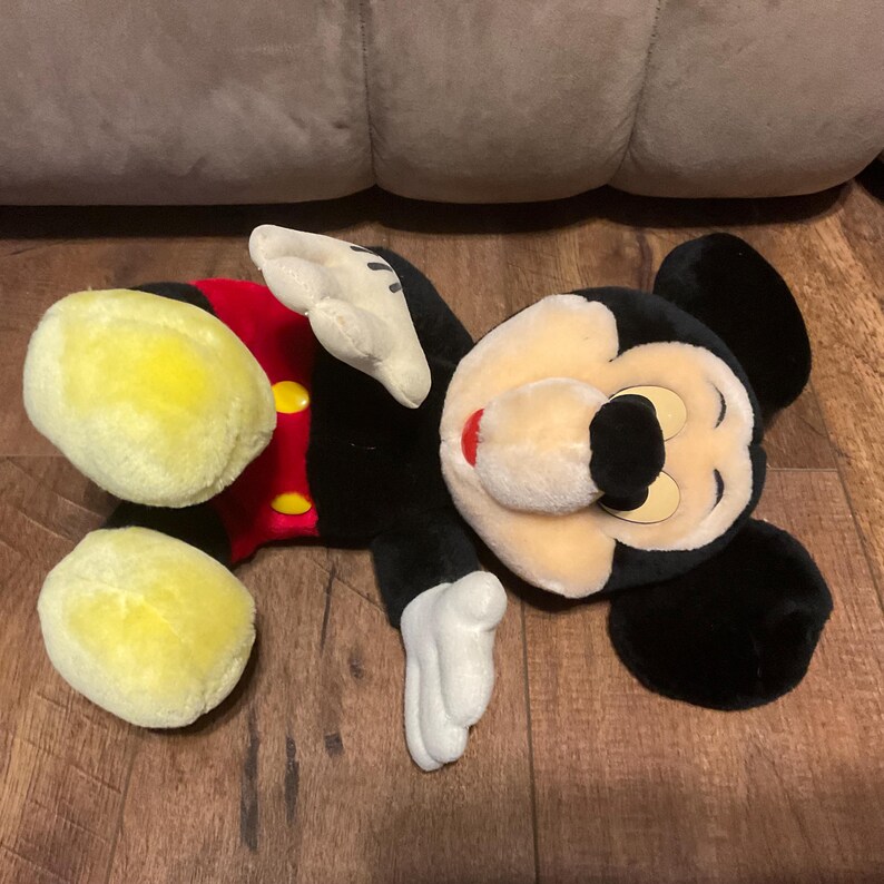 Pu&ograve; includere: Un peluche di Topolino, sdraiato su una superficie di legno. Il giocattolo presenta un corpo nero, grandi piedi gialli, guanti bianchi e una camicia rossa con bottoni gialli. Il viso &egrave; color crema con un naso nero e un'espressione sorridente.