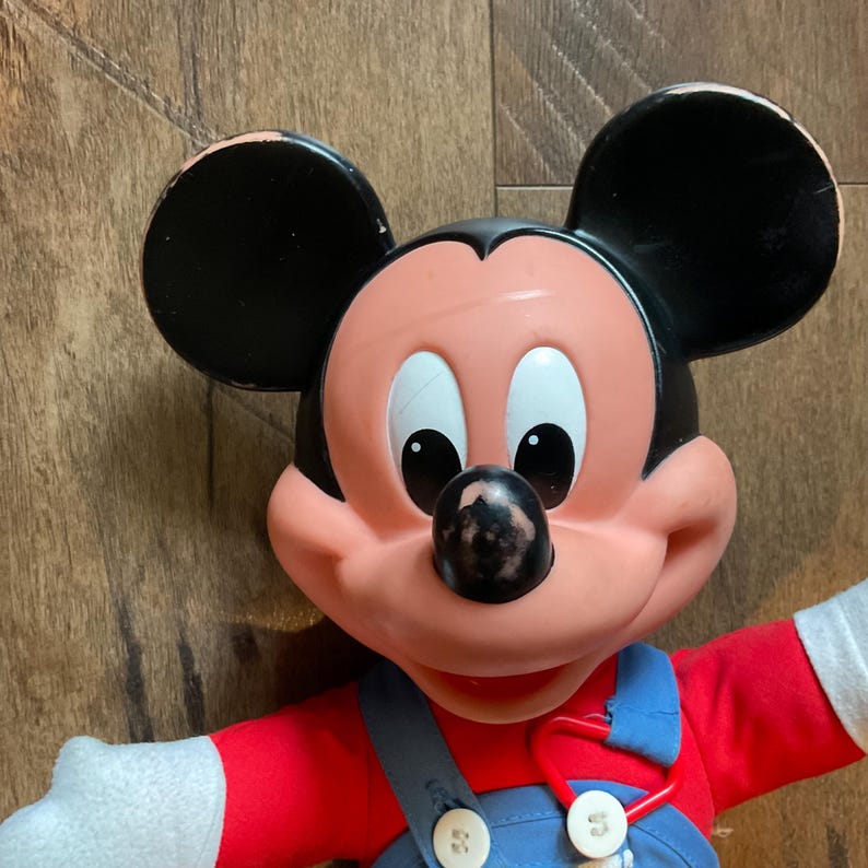 May include: A vintage Mickey Mouse plush doll. The doll has a black head and ears, a pink face, and a red shirt. It is wearing blue overalls with white buttons and white gloves. The doll's nose has some wear.