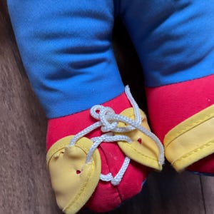 May include: Close-up of a doll's legs and feet. The doll's legs are blue, and the feet are red with yellow accents. White laces are tied in a bow over the yellow shoe details. The doll is on a wooden surface.