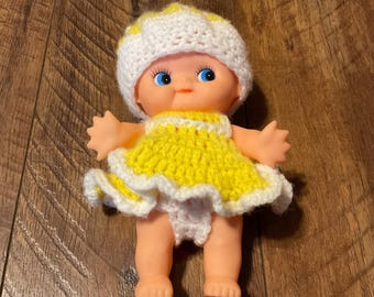 Könnte beinhalten: Vintage Babypuppe mit gelbem und weißem gehäkeltem Kleid und Hut. Die Puppe hat blaue Augen und besteht aus einem weichen, gummiartigen Material. Das Kleid hat einen Rüschensaum.