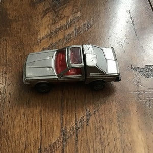 May include: A silver toy pickup truck with a red interior and a clear sunroof. The toy truck has black wheels and is set against a wooden background.
