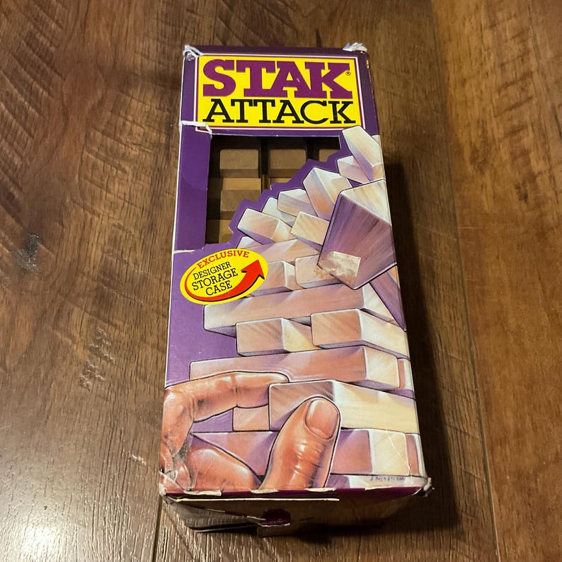 Peut inclure: Bo&icirc;te de jeu Stak Attack avec un design violet et jaune. La bo&icirc;te pr&eacute;sente le nom du jeu et une illustration de blocs de bois. Un &eacute;tui de rangement design exclusif est &eacute;galement pr&eacute;sent&eacute; sur la bo&icirc;te.