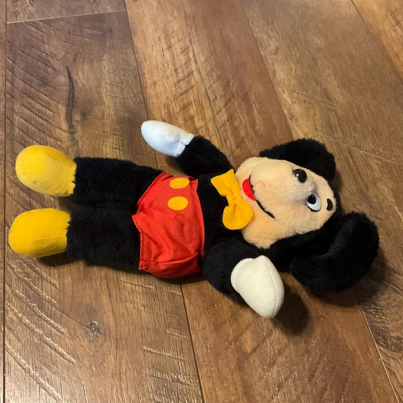 K&ouml;nnte beinhalten: Ein Stofftier von Micky Maus, liegend auf einer Holzoberfl&auml;che. Das Spielzeug hat einen schwarzen K&ouml;rper, wei&szlig;e Handschuhe, gelbe Schuhe und rote Shorts mit gelben Kn&ouml;pfen. Es hat ein cremefarbenes Gesicht, schwarze Ohren und eine gelbe Fliege.
