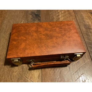 May include: A brown leather-bound game box with a brass handle and clasps. The box has a textured surface and is closed.