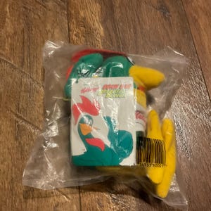 May include: A green and yellow rooster plush toy with a red comb and wattle. The toy is packaged in a clear plastic bag with a Kellogg's 'Tony the Tiger's Breakfast Bunch' tag attached. The tag is white with red, yellow, and blue accents.