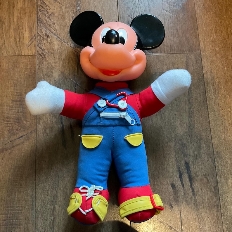May include: A vintage Mickey Mouse plush doll. The doll has a peach-coloured face, black ears, and a red shirt. It wears blue dungarees with yellow pockets and red and yellow shoes. The doll's arms have white gloves.