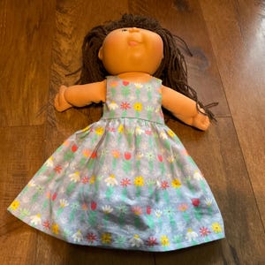 May include: A vintage doll with brown yarn hair and a light skin tone. The doll is wearing a light blue dress with a floral pattern of white, yellow, and red flowers. The doll is lying on a wooden surface.