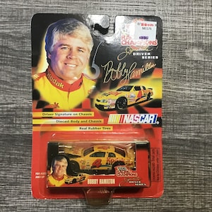 May include: A NASCAR Racing Champions diecast car of Bobby Hamilton, packaged on a red and black card. The card features a photo of Bobby Hamilton and his signature. The car is yellow with the number 4.