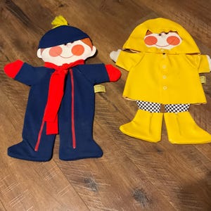 May include: Two vintage plush dolls. One doll is dressed in a navy blue outfit with a red scarf and red accents. The other doll is wearing a yellow raincoat, boots, and checkered shorts. Both dolls have smiling faces.