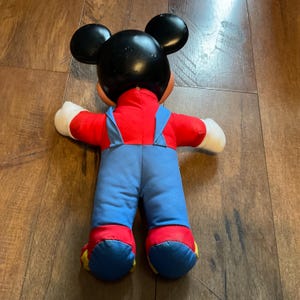 May include: A vintage Mickey Mouse soft toy, seen from behind. The toy has a black head with large ears, a red shirt, blue dungarees, and yellow and red shoes. The arms are white, and the toy is on a wooden surface.