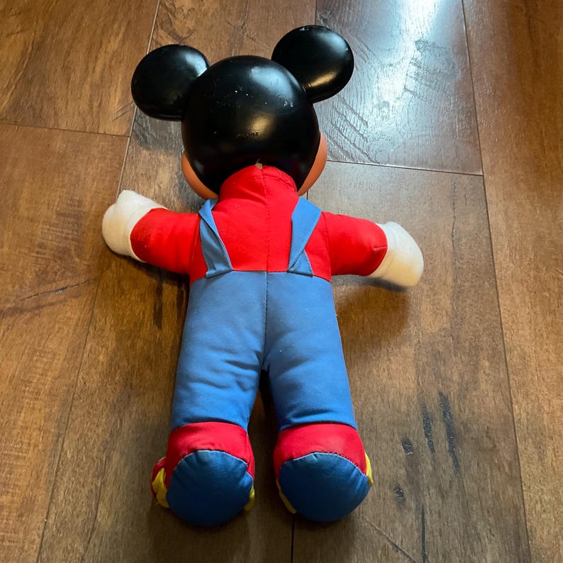 May include: A vintage Mickey Mouse soft toy, seen from behind. The toy has a black head with large ears, a red shirt, blue dungarees, and yellow and red shoes. The arms are white, and the toy is on a wooden surface.