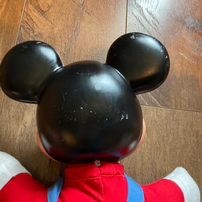 May include: A vintage Mickey Mouse plush toy, seen from the back. The toy has a black head with large ears, a red shirt, blue overalls, and white gloves. The plush is set against a wooden background.