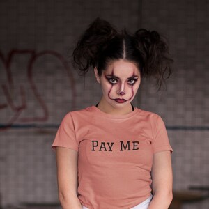 May include: A coral-colored t-shirt with the text "PAY ME" in black. The person is wearing white shorts, black knee-high socks, and white sneakers, holding a baseball bat. The background is an industrial setting.