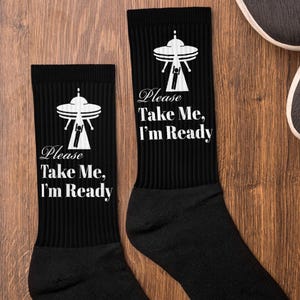 May include: A pair of black crew socks with a white alien spaceship graphic and the text "Please Take Me, I'm Ready".