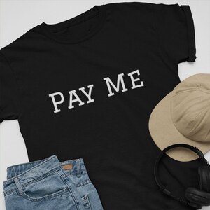 May include: Black t-shirt with the text "PAY ME" in white, paired with denim shorts, black sneakers, a tan baseball cap, headphones, and a skateboard. The items are arranged on a white surface.