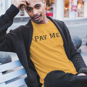 May include: Mustard yellow t-shirt with the text "PAY ME" in black, worn by a person. The person is wearing a dark gray cardigan and black pants. The person is sitting on a light blue bench.