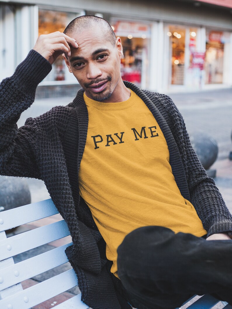 May include: Mustard yellow t-shirt with the text "PAY ME" in black, worn by a person. The person is wearing a dark gray cardigan and black pants. The person is sitting on a light blue bench.