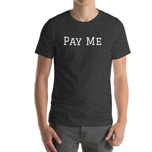 May include: Dark gray t-shirt with the white text "PAY ME" across the chest. The shirt is a crew neck style and appears to be made of a soft, comfortable material. The model is wearing gray pants.