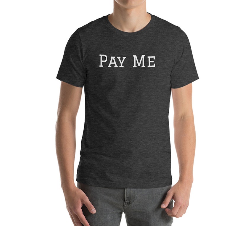 May include: Dark gray t-shirt with the white text "PAY ME" across the chest. The shirt is a crew neck style and appears to be made of a soft, comfortable material. The model is wearing gray pants.