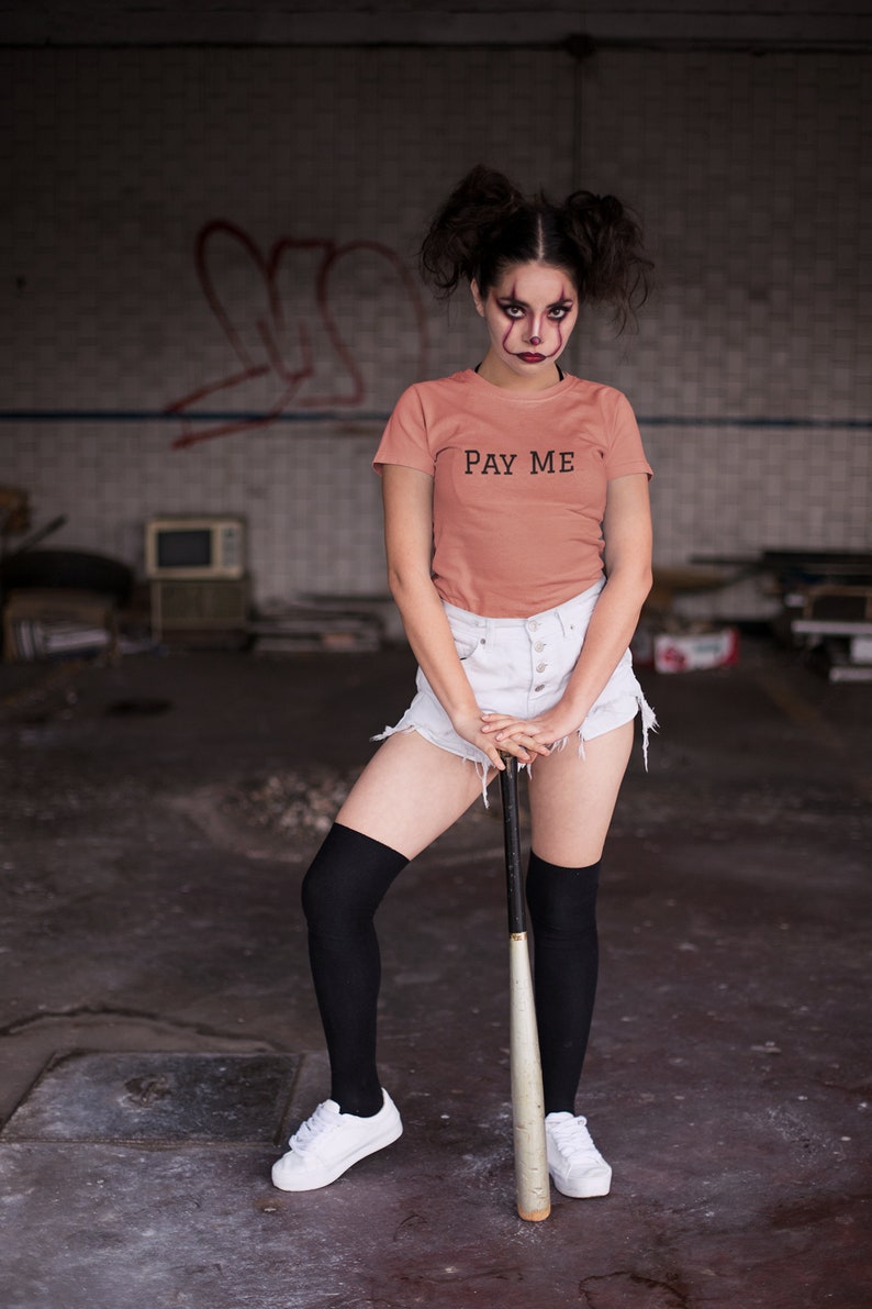 May include: A coral-colored t-shirt with the text "PAY ME" in black. The person is wearing white shorts, black knee-high socks, and white sneakers, holding a baseball bat. The background is an industrial setting.