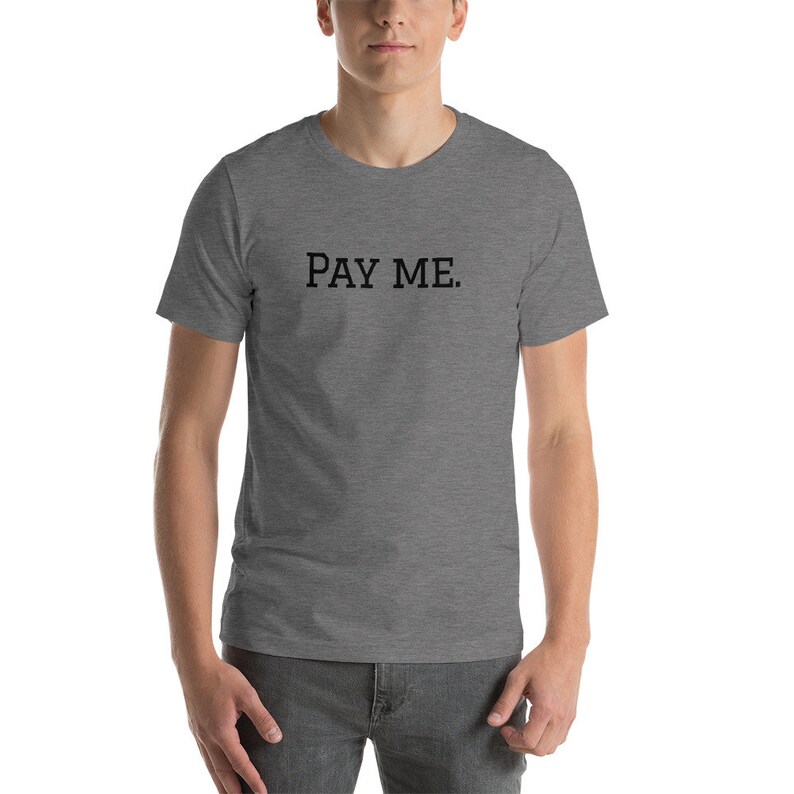 May include: A heather gray t-shirt with the text "PAY ME." printed in black. The shirt is short-sleeved and appears to be made of a soft material. The person wearing the shirt is standing in front of a neutral background.