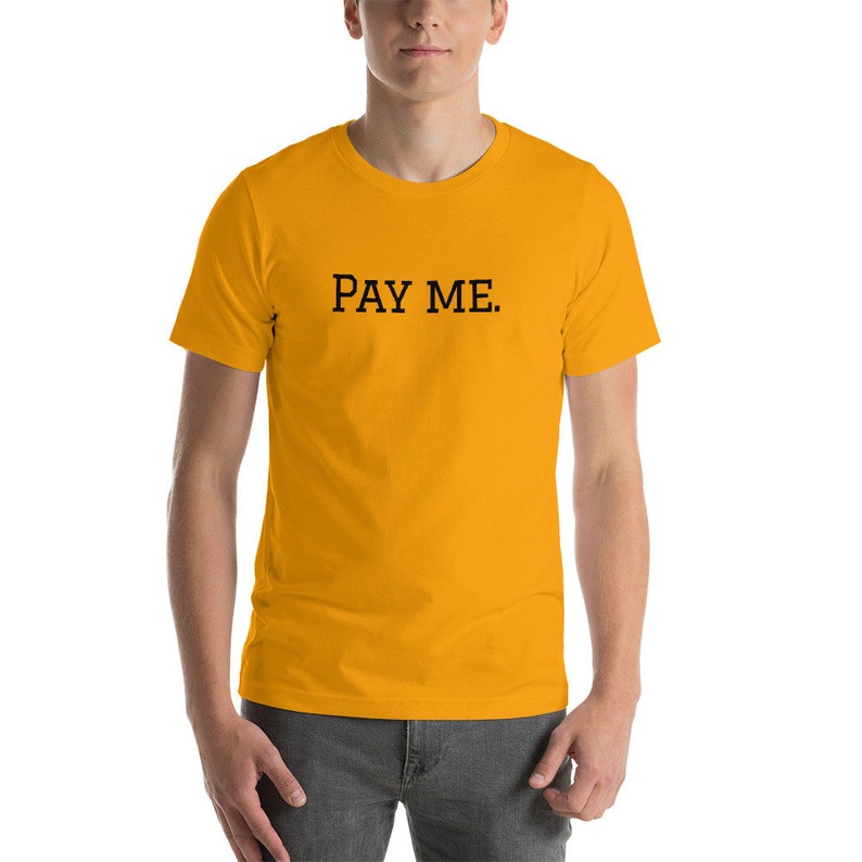 May include: Mustard yellow t-shirt with the text "PAY ME" printed in black. The short-sleeved shirt is made of a soft material and is being worn.
