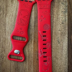 May include: Red watch bands with travel-themed designs. One band features an airplane window, clouds, and hearts. The other band has a gate sign, hearts, a chair, a passport, and a suitcase. The bands are made of a flexible material.