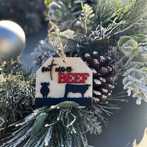 May include: A wooden Christmas ornament with a brown background and the text "Eat More Beef" in black and red lettering. The ornament features a silhouette of a cow and a bull.