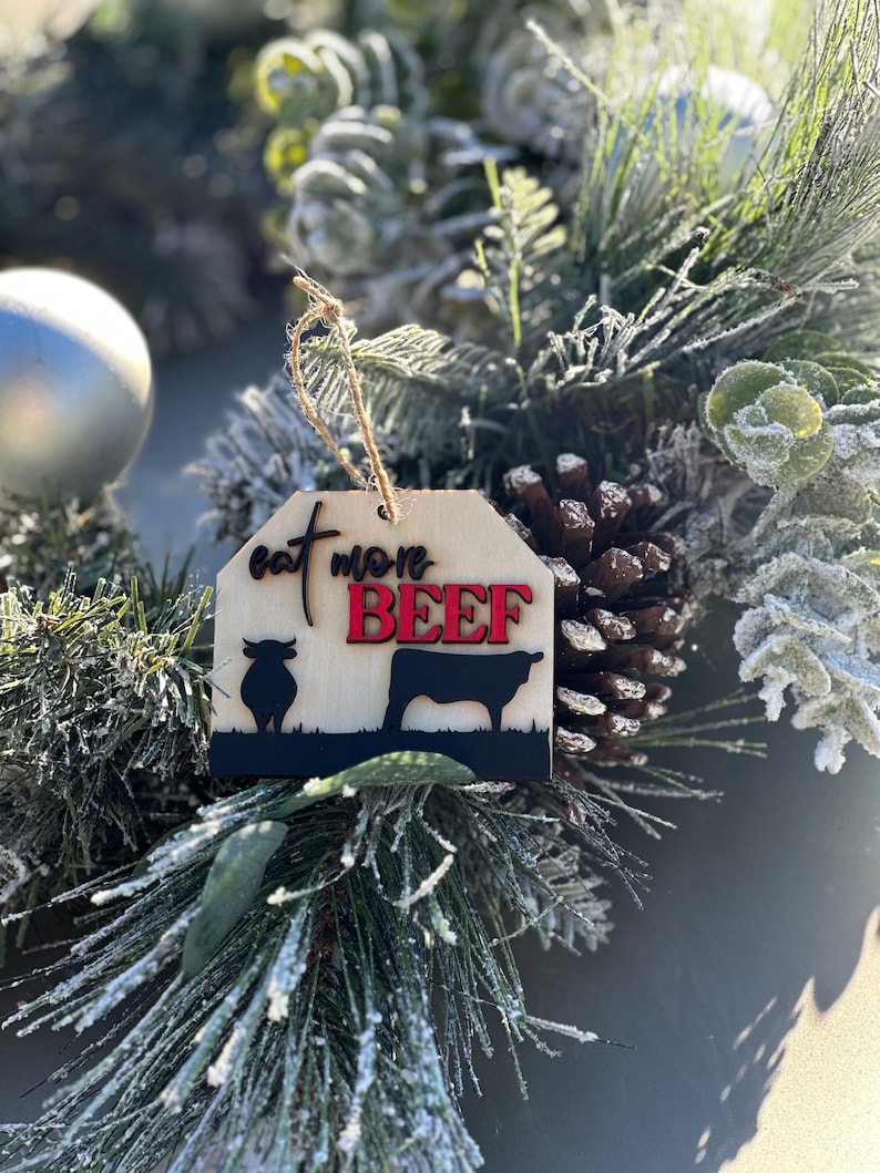 May include: A wooden Christmas ornament with a brown background and the text "Eat More Beef" in black and red lettering. The ornament features a silhouette of a cow and a bull.