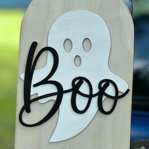 May include: A wooden sign with a white ghost cutout and the word "Boo" in black lettering.