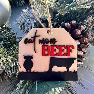 May include: Wooden Christmas ornament with a brown background and the text "Eat More Beef" in black and red lettering. The ornament features two black silhouettes of cows.