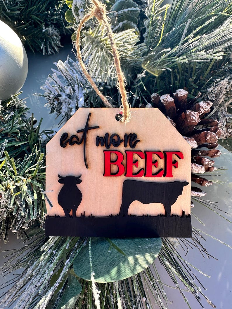 May include: Wooden Christmas ornament with a brown background and the text "Eat More Beef" in black and red lettering. The ornament features two black silhouettes of cows.