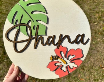 May include: A round wooden sign with the name "Dhana" written in brown cursive lettering. The sign is decorated with a green tropical leaf and a pink hibiscus flower.
