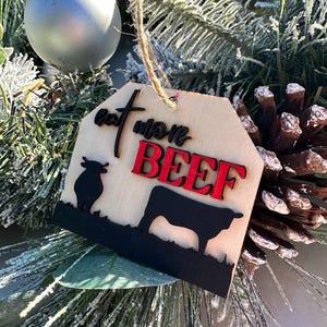 May include: A wooden Christmas ornament with a brown background and black silhouette of a cow. The ornament has the text "Eat More Beef" in red and black lettering.