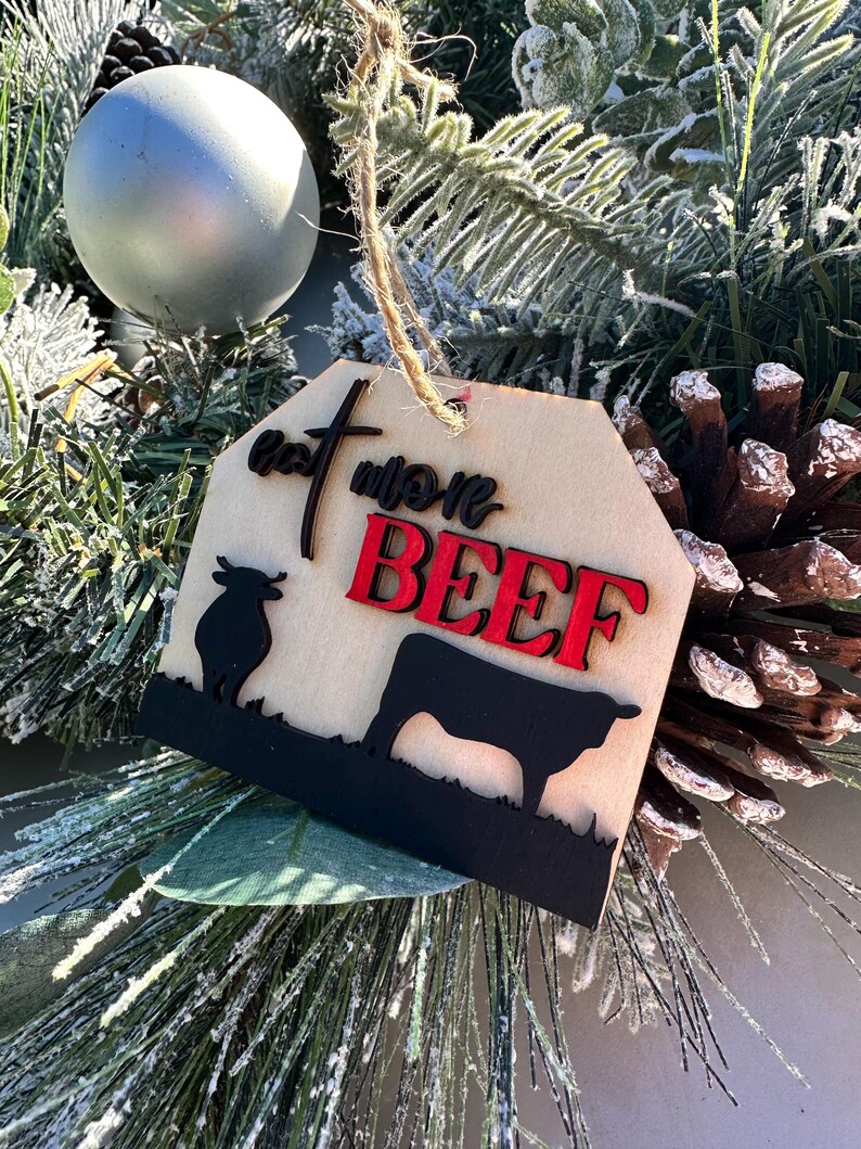 May include: A wooden Christmas ornament with a brown background and black silhouette of a cow. The ornament has the text "Eat More Beef" in red and black lettering.