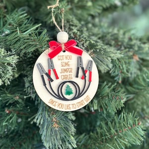 May include: A wooden Christmas ornament with the text "GOT YOU SOME JUMPER CABLES SINCE YOU LIKE TO START SHIT." The ornament features jumper cables and a small green Christmas tree. A red bow and wooden bead are at the top.