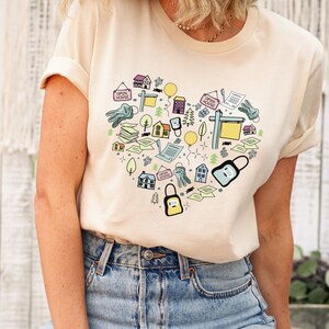 Puede incluir: Una camiseta de color crema con un diseño en forma de corazón que presenta varias ilustraciones relacionadas con bienes raíces. El diseño incluye casas, llaves, letreros y otras imágenes relacionadas.