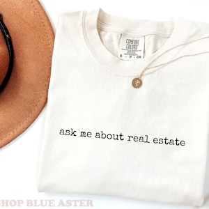 May include: Ivory-colored t-shirt with the text "ask me about real estate" in black. A gold necklace with a pendant is draped on the shirt. A brown hat and dried plant are in the background. The shirt is from Comfort Colors.