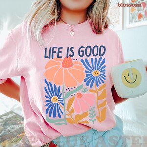 K&ouml;nnte beinhalten: Hellrosa T-Shirt mit der Aufschrift "LIFE IS GOOD" &uuml;ber einem Blumendesign in Orange, Blau und Rosa. Eine wei&szlig;e Tasse mit einem gelben Smiley wird in der Hand gehalten.