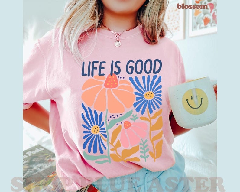 K&ouml;nnte beinhalten: Hellrosa T-Shirt mit der Aufschrift "LIFE IS GOOD" &uuml;ber einem Blumendesign in Orange, Blau und Rosa. Eine wei&szlig;e Tasse mit einem gelben Smiley wird in der Hand gehalten.