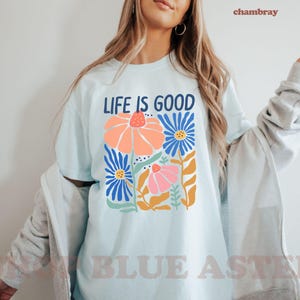 K&ouml;nnte beinhalten: Hellblaues T-Shirt mit dem Text "LIFE IS GOOD" &uuml;ber einem Blumendesign. Das Design zeigt orange, blaue und rosa Blumen mit gr&uuml;nen und gelben Akzenten. Das Shirt hat einen l&auml;ssigen Stil.