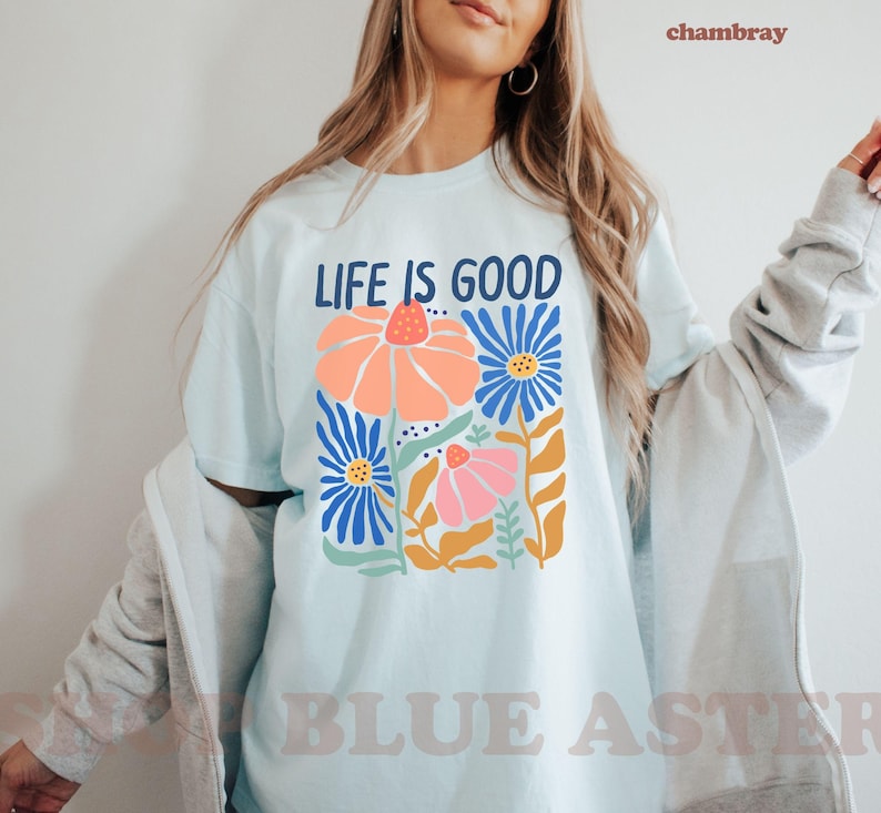 K&ouml;nnte beinhalten: Hellblaues T-Shirt mit dem Text "LIFE IS GOOD" &uuml;ber einem Blumendesign. Das Design zeigt orange, blaue und rosa Blumen mit gr&uuml;nen und gelben Akzenten. Das Shirt hat einen l&auml;ssigen Stil.