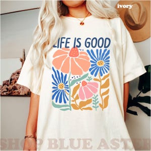 K&ouml;nnte beinhalten: Elfenbeinfarbenes T-Shirt mit dem Text "LIFE IS GOOD" in Marineblau. Das Shirt zeigt ein Blumendesign mit orangefarbenen, blauen und rosa Blumen. Das Design umfasst gr&uuml;ne Bl&auml;tter und Stiele.