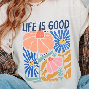 K&ouml;nnte beinhalten: Elfenbeinfarbenes T-Shirt mit dem Text "LIFE IS GOOD" in Marineblau. Das Shirt zeigt ein Blumendesign mit orangefarbenen, blauen und rosa Blumen. Die Blumen haben gelbe und gr&uuml;ne Stiele und Bl&auml;tter. Ein l&auml;ssiges Kleidungsst&uuml;ck f&uuml;r den Alltag.