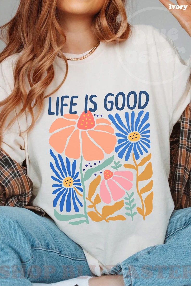 K&ouml;nnte beinhalten: Elfenbeinfarbenes T-Shirt mit dem Text "LIFE IS GOOD" in Marineblau. Das Shirt zeigt ein Blumendesign mit orangefarbenen, blauen und rosa Blumen. Die Blumen haben gelbe und gr&uuml;ne Stiele und Bl&auml;tter. Ein l&auml;ssiges Kleidungsst&uuml;ck f&uuml;r den Alltag.