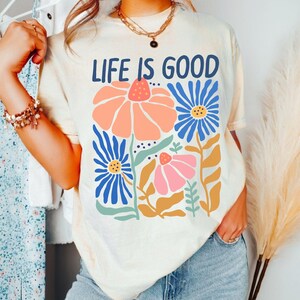 K&ouml;nnte beinhalten: Cremefarbenes T-Shirt mit dem Schriftzug "LIFE IS GOOD" in Marineblau. Das Shirt zeigt ein Blumendesign mit orangefarbenen, rosa und blauen Blumen. Das Model tr&auml;gt eine blaue Jeans und eine goldene Halskette.