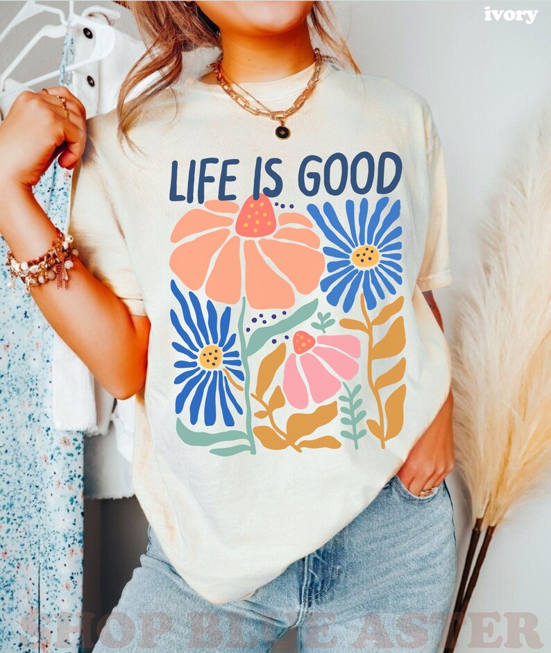 K&ouml;nnte beinhalten: Cremefarbenes T-Shirt mit dem Schriftzug "LIFE IS GOOD" in Marineblau. Das Shirt zeigt ein Blumendesign mit orangefarbenen, rosa und blauen Blumen. Das Model tr&auml;gt eine blaue Jeans und eine goldene Halskette.